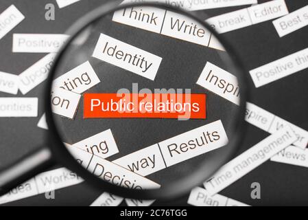 Magnifier glass over the red inscription public relations cut out of paper. Surrounded by other inscriptions on a dark background. Word cloud concept. Stock Photo
