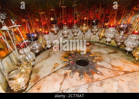 The Grotto of the Nativity, a rectangular alcove beneath the church, is the Church of the Nativity's focal point. Entered by a flight of steps by the church altar, this is the cave that has been honored as the site of Christ's birth since at least the 2nd century. A silver star on the floor marks the very spot where Christ is believed to have been born. Many people believe rubbing the star with their hands or objects blesses them and the objects. The floor is paved in marble, and 15 lamps hang above the star. The Church of the Nativity in Bethlehem, West Bank, Israel,  is a major Christian hol Stock Photo