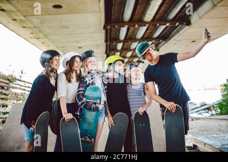 group of skateboarders children make social media content on phone while spending time on ramp in skate park. Friends stand together with skate boards Stock Photo