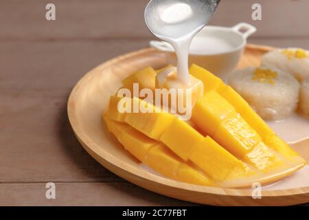 Fresh yellow mango peel and Sliced cubes with sticky rice. Spoon pouring coconut milk on top mango. Favorite thai dessert summer. Mango on wood plate. Stock Photo