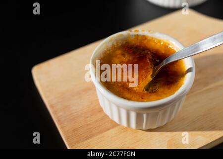 Homemade creme brulee in white ramekin  with sugar burn top up cracking with spoon on black background with copy space. French dessert creme brulee Stock Photo