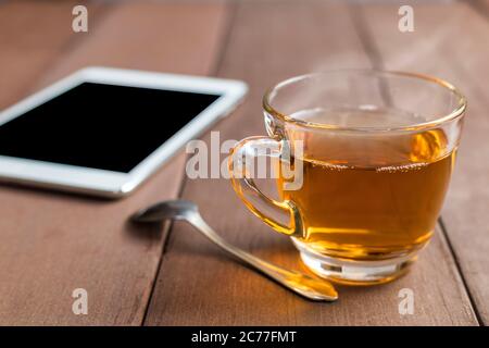Close up hot tea cup on wood table with tea spoon and tablet shot on morning time with sun ray background. Soft focus and selective focus to tea cup. Stock Photo