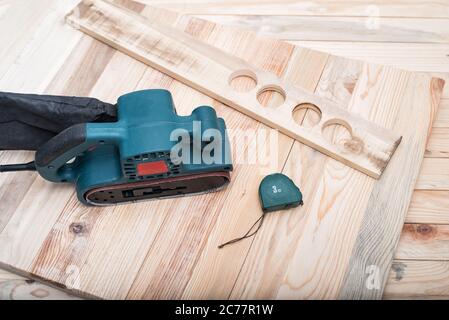 Electric belt sander, measuring tape and workpiece lying on a light brown wooden table. Woodworking, sanding machine Stock Photo