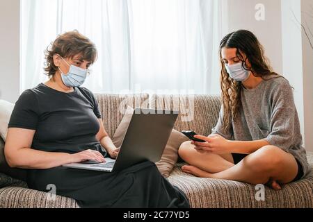 Mother and daughter protected with a mask against coronavirus, covid-19 or any other disease, sitting in their living room using a phone and a compute Stock Photo
