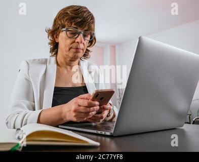 Old lady using her smartphone while working on her laptop. Business woman working from home. Senior woman working from home. Female entrepreneur works Stock Photo