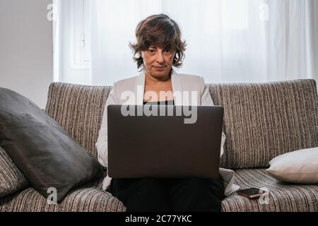 Business woman using her laptop at home. Business woman working from home. Woman on the couch. Older woman working. Old lady using her laptop. Working Stock Photo