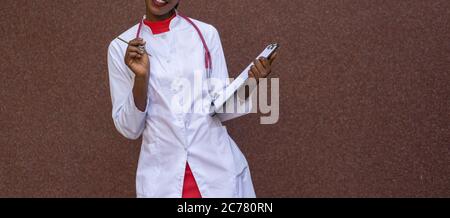 black young woman doctor, in a white coat, with a phonendoscope, holds a folder for medical papers in a rue, against the background of a hospital. Stock Photo