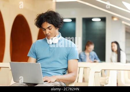 Concentrated dark-skinned student guy sitting on chair in library and using laptop while working on university project Stock Photo