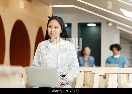 Content Asian student in headscarf sitting on chair in auditorium and using laptop at lecture Stock Photo