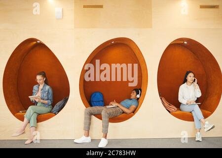 Modern multi-ethnic students in casual outfits sitting in egg-shaped wall chairs and using gadgets during break Stock Photo