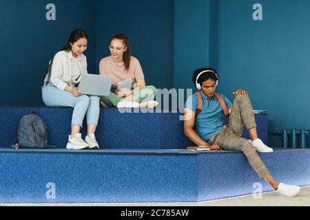 Multi-ethnic university students sitting on steps and using workbooks and devices while doing task before class in blue lobby Stock Photo