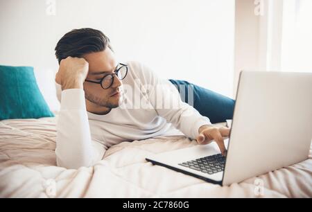 Tired caucasian man with beard and eyeglasses chatting on laptop in bed Stock Photo