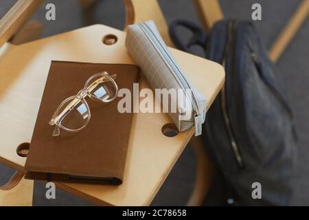 Close-up of checkered pencil case, brown notepad and eyeglasses on wooden chair in university room Stock Photo