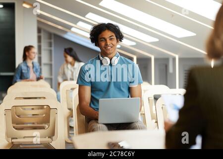 Smiling curious Mixed race guy with wireless headphones around neck sitting with laptop on chair and talking to university teacher Stock Photo