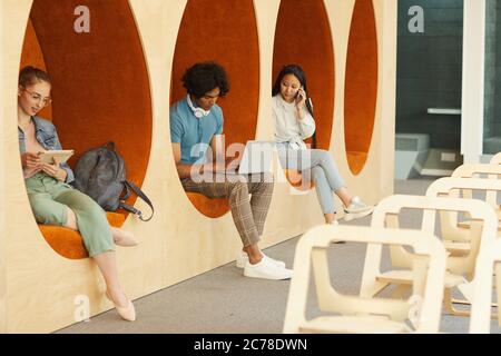 Modern multi-ethnic students sitting inside of wall chairs in university hall and using devices for education Stock Photo