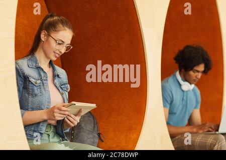 Young multi-ethnic students sitting in comfortable wall chairs and using devices while preparing for seminar during break Stock Photo