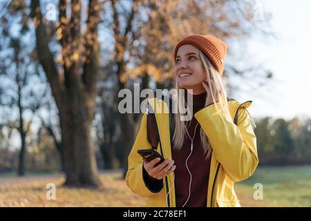 Beautiful smiling woman wearing yellow raincoat listening music looking away outdoors. Young emotional tourist walking in park. Autumn travel Stock Photo