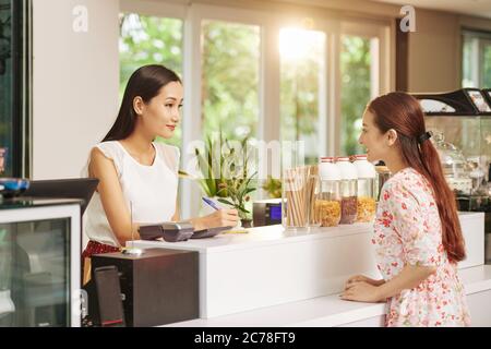 Smiling young female barista taking notes when talking to customer ordering coffee and breakfast Stock Photo