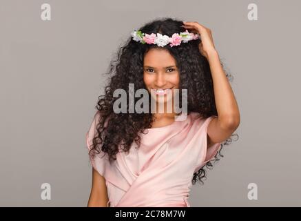 Pretty young Afro American bridesmaid in a pink dress holding a coronet of flowers to her long curly hair with a happy charming smile against a beige Stock Photo