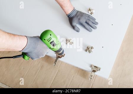 Furniture assembly using drill. Male hands in gray gloves, master collects furniture using tools instrument drill in house. Moving, home improvement Stock Photo