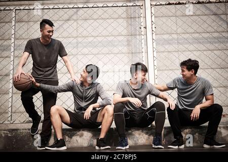 group of four asian young adult men resting relaxing talking chatting on outdoor basketball court Stock Photo