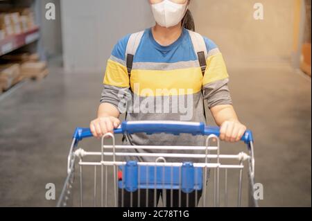 The asian woman wearing surgical mask doing shopping and pushing with her cart in cargo or warehouse.  Prepare shopping the furniture interior concept Stock Photo