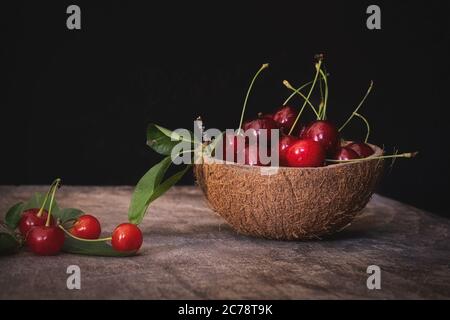 Fresh cherries in a coconut shell bowl on a wooden table on black background. Healthy eating. Stock Photo