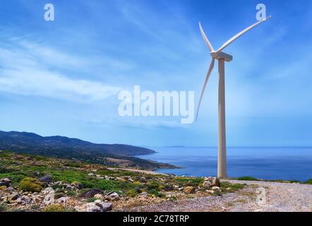 Single windmill turbine on hilltop of seashore in colorful landscape against dynamic blue sky with clouds and winding road. Stock Photo