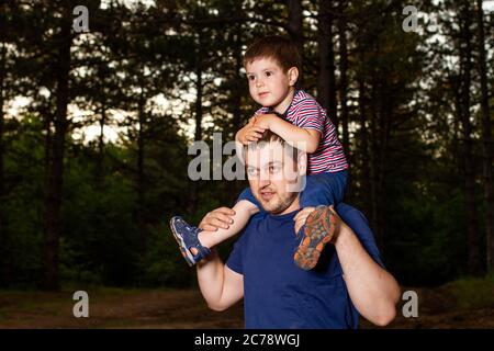 Dad carries his son on his shoulders around his neck. Father and son walk in the woods in the wild among the trees in the evening. Love and family Stock Photo