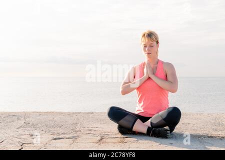 beautiful blonde woman practicing yoga at dawn sitting in front of the sea, concept of mental health and relaxation, copy space for text Stock Photo