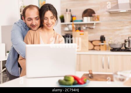 Boyfriend hugs girlfriend in kitchen while using laptop. Happy loving cheerful romantic in love couple at home using modern wifi wireless internet technology Stock Photo