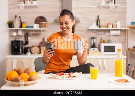 Amused young woman reading funny news on phone in the kitchen drinking a cup of coffee. Housewife using modern tehcnology and drinking healthy, natural, homemade coffee. Refreshing morning Stock Photo