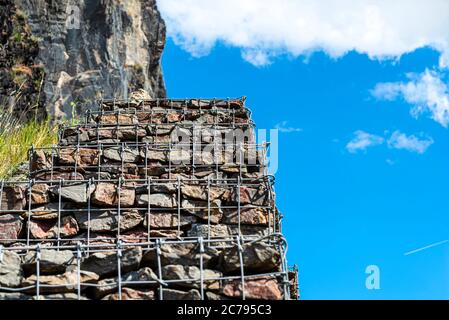 A stone wall standing by the road, made of stones protected by metal, galvanized mesh in the background of the blue sky with clouds, selective focus. Stock Photo