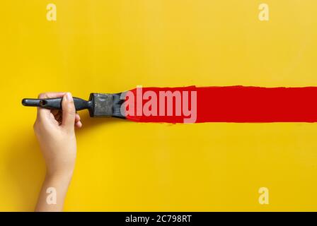 hand holding paint brush, painted red color isolated on yellow background with copy space. home improvement concept Stock Photo
