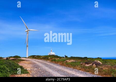 Single windmill turbine, small traditional Greek Orthodox church, road. Colorful landscape on hilltop at blue sky and clouds on sunny summer day. Stock Photo
