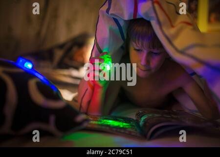 Boy with green flashlight reading magazine under blanket Stock Photo