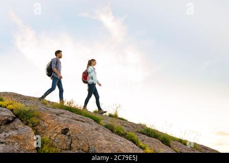 Couple of Young Happy Travelers Hiking with Backpacks on the Rocky Trail at the Evening. Family Travel and Adventure Stock Photo