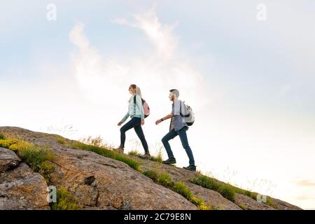 Couple of Young Happy Travelers Hiking with Backpacks on the Rocky Trail at the Evening. Family Travel and Adventure Stock Photo