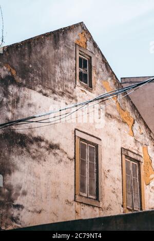 Side vertical shot of an old flaked facade of an antique residential house with a triangle roof and three windows; stretched wires in the foreground Stock Photo