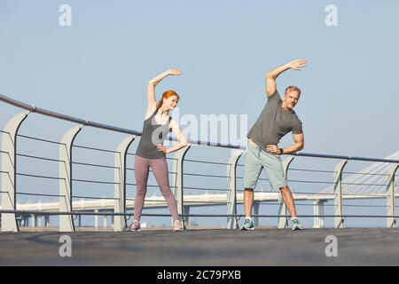 Athletic mature couple standing on city embankment and doing side bend together before training Stock Photo