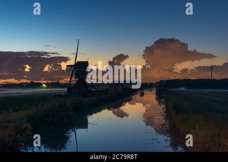 Night image of a Dutch windmill along the water of a canal. Layer of fog in the fields and dramatic clouds in the background. Stock Photo