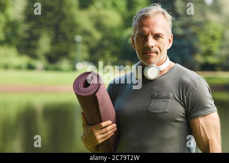 Portrait of handsome mature man with wireless headphones around neck holding rolled fitness mat in city park Stock Photo