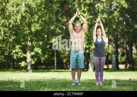 Serious sporty mature couple standing with raised hands in Namaste and doing breath exercise in summer park Stock Photo