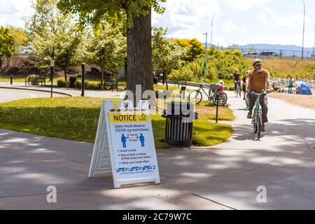 Kelowna, BC / Canada - May 18, 2020: Sign Advocating Social Distancing During Covid-19 Pandemic As Cyclist Rides By and Citizens Enjoy The Park. Stock Photo