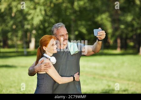 Handsome mature sportsman with towel on shoulder embracing wife and photographing with her after training in park Stock Photo