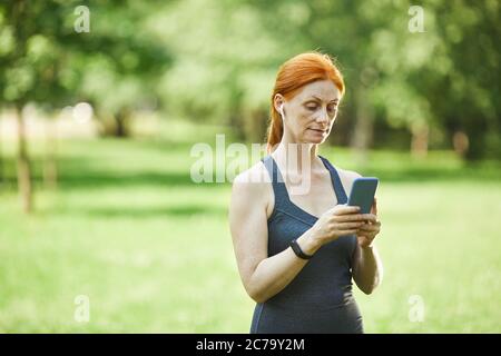 Redhead mature woman in wireless ear buds standing in park and choosing music for fitness using smartphone Stock Photo