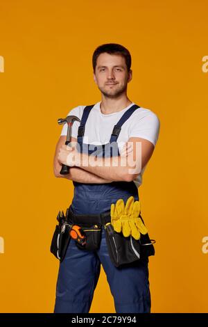 Young caucasian repairman worker in yellow hard hat and uniform holding hammer. handyman wearing tool belt isolated on orange studio background Stock Photo