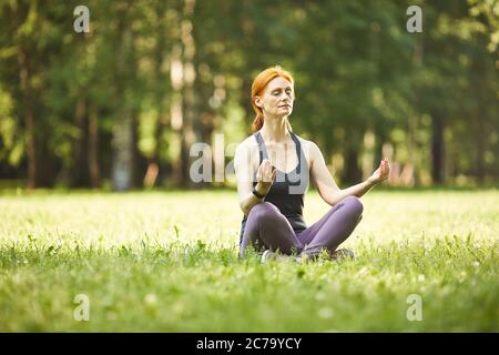 Serene relaxed mature redhead woman sitting with crossed legs on grass and meditating with closed eyes in park Stock Photo