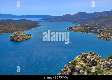 View of harbor between Spinalonga island and peninsula, known as Kalydon, and Plaka village from above and rocky slope in foreground, Crete, Greece. Stock Photo