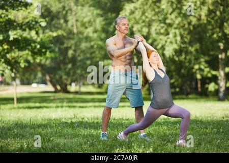 Professional muscle-bound trainer adjusting yoga pose of woman while they practicing in summer park Stock Photo